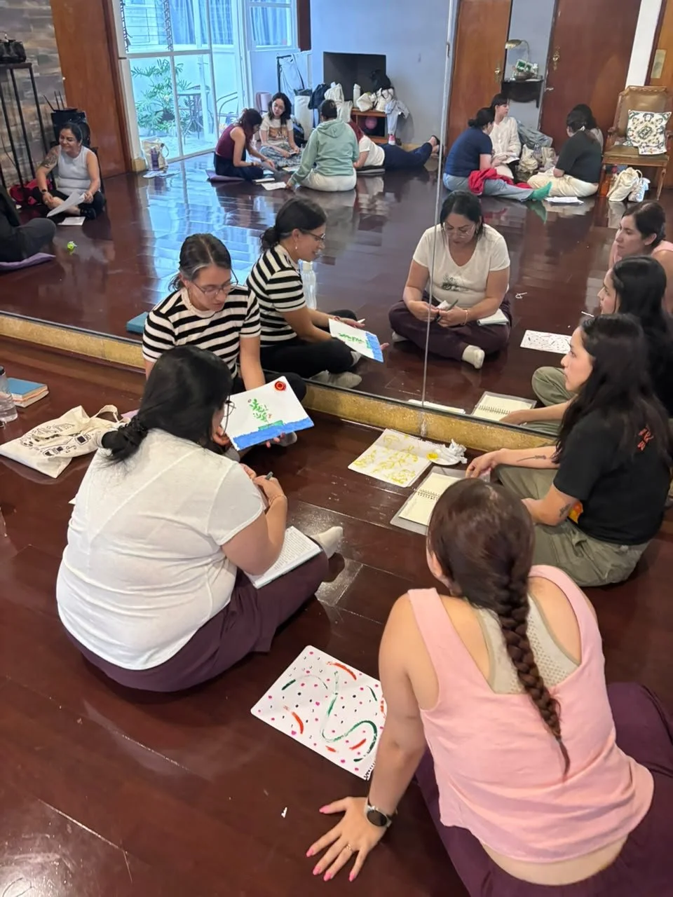 Grupo de personas sentadas en el suelo, participando en un taller o reunión, con materiales de arte y libros alrededor, en una habitación con una pared de espejo y una ventana.