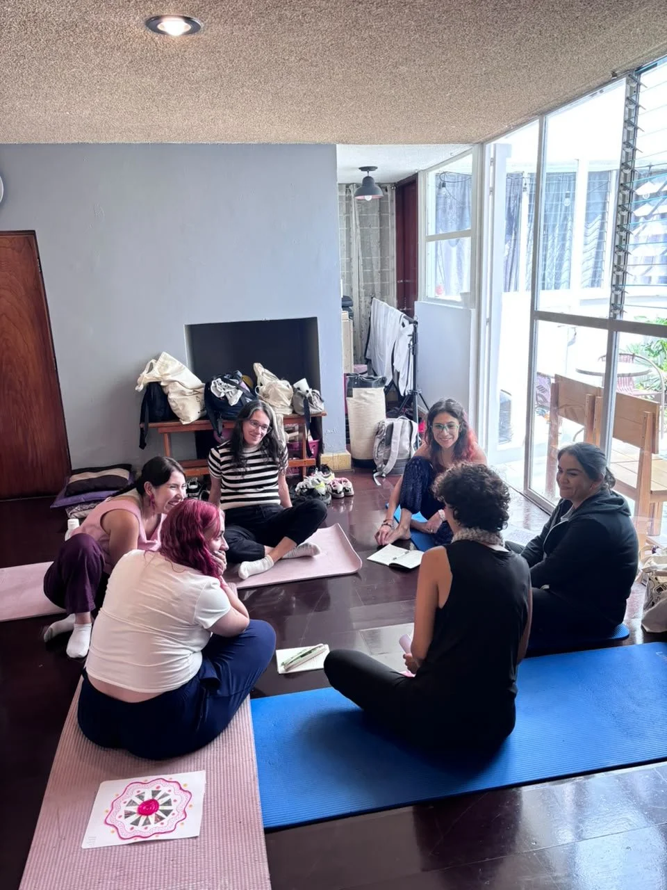 Grupo de seis mujeres sentadas en un piso de madera, participando en una sesión de yoga o meditación, con luz natural entrando por ventanas grandes.