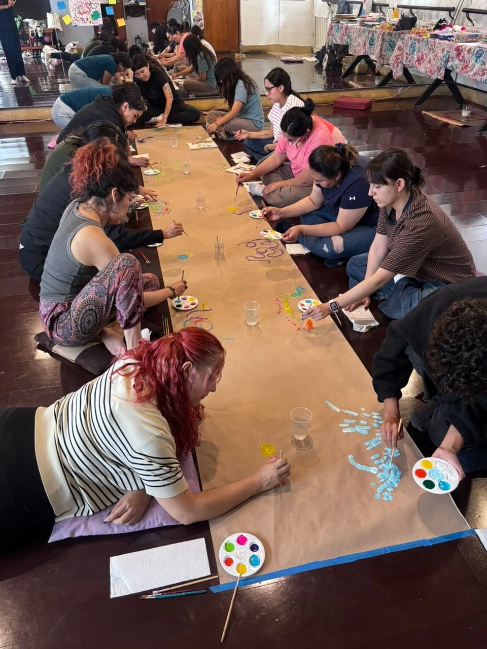 Varias personas adultas participan en una actividad de pintura y arte en un piso de madera, sentadas alrededor de una mesa larga cubierta con papel kraft, usando pinceles y pinturas de colores. En el fondo, hay espejos, mesas con objetos y decoracion