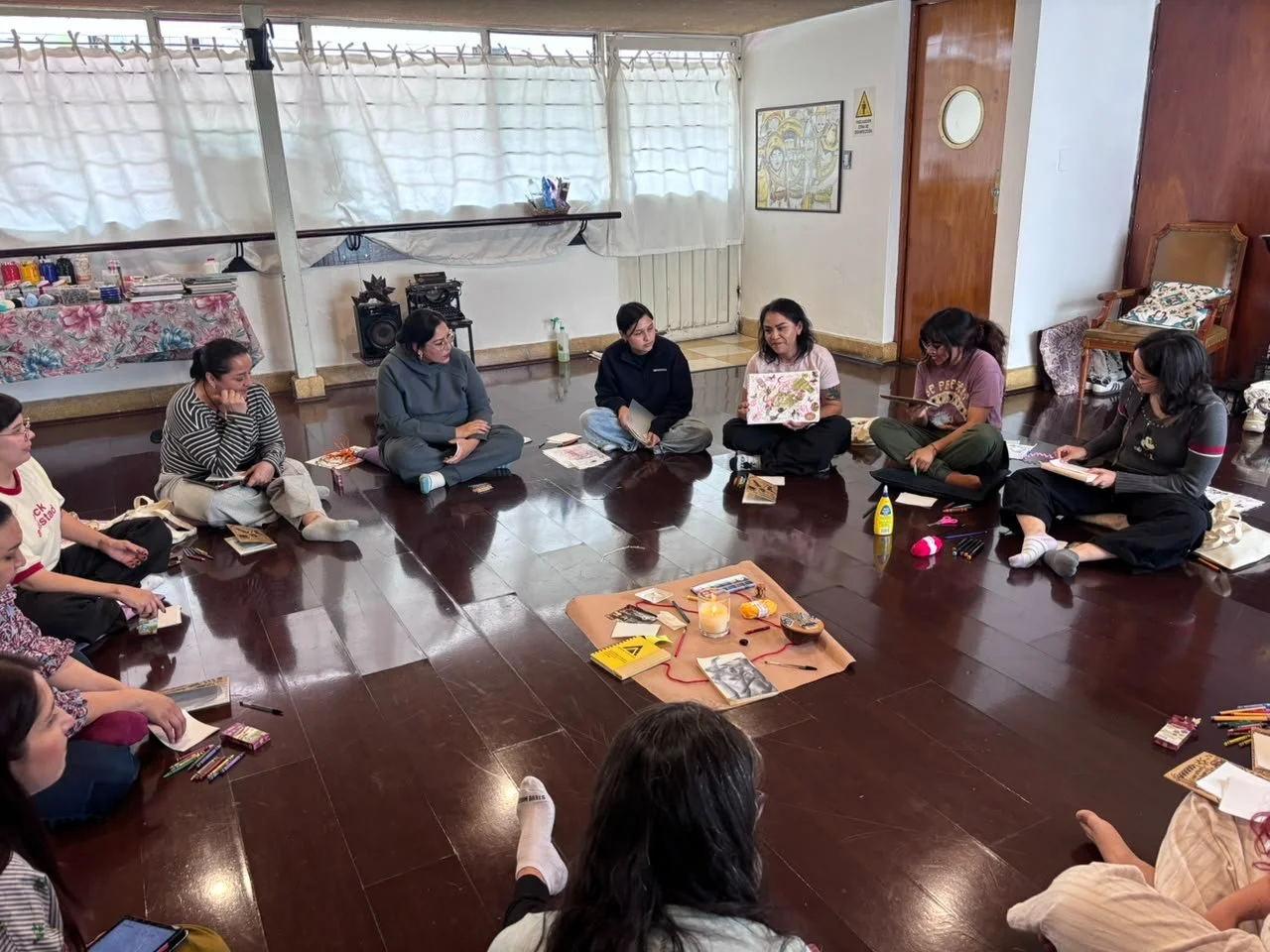 Grupo de mujeres sentadas en círculo en un taller o reunión, en un espacio interior con piso de madera y paredes blancas, con una mesa con materiales y decoraciones en el centro.