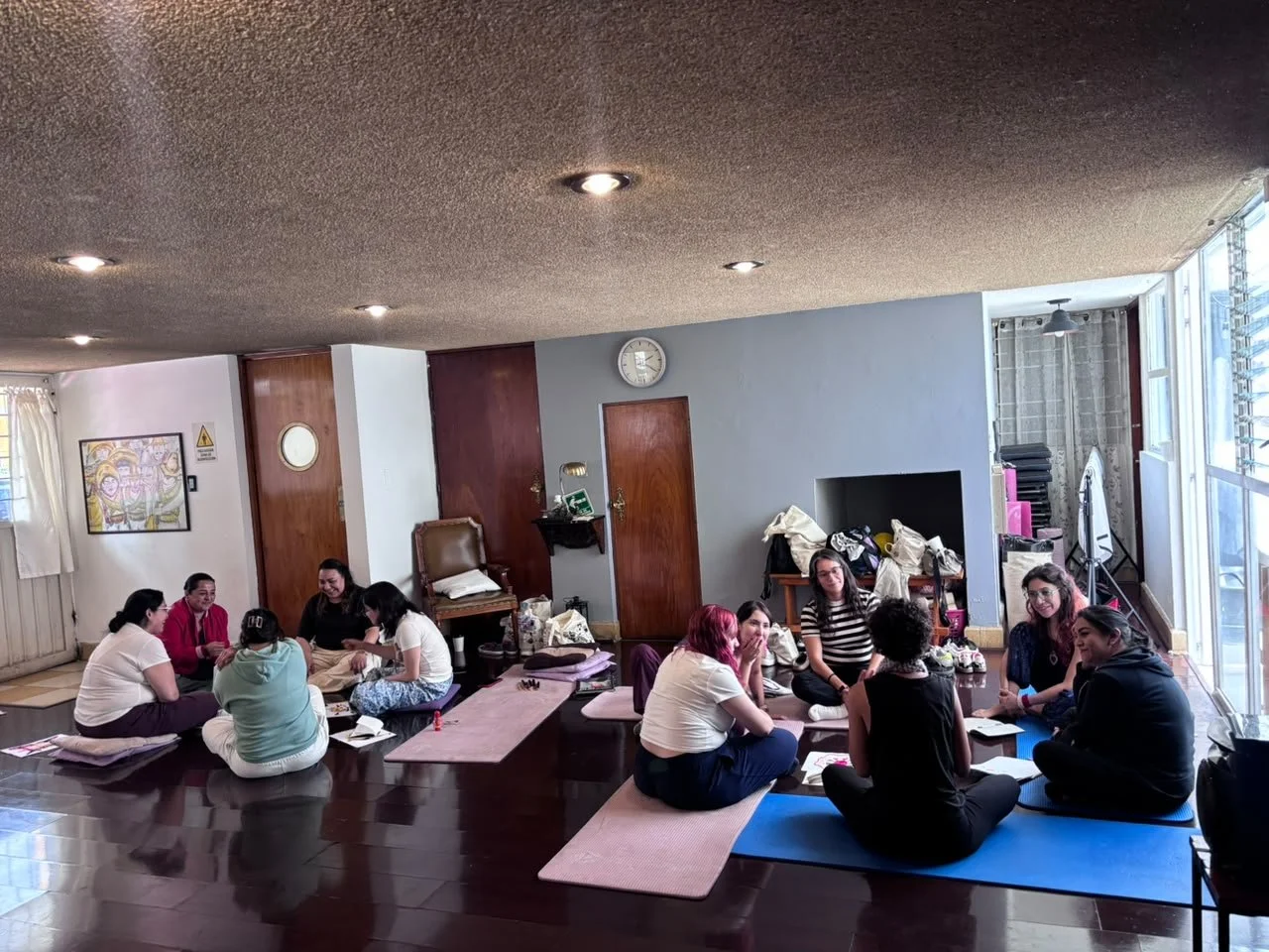 Grupo de personas participando en una clase en un salón con ventanas grandes y pisos de madera oscura.