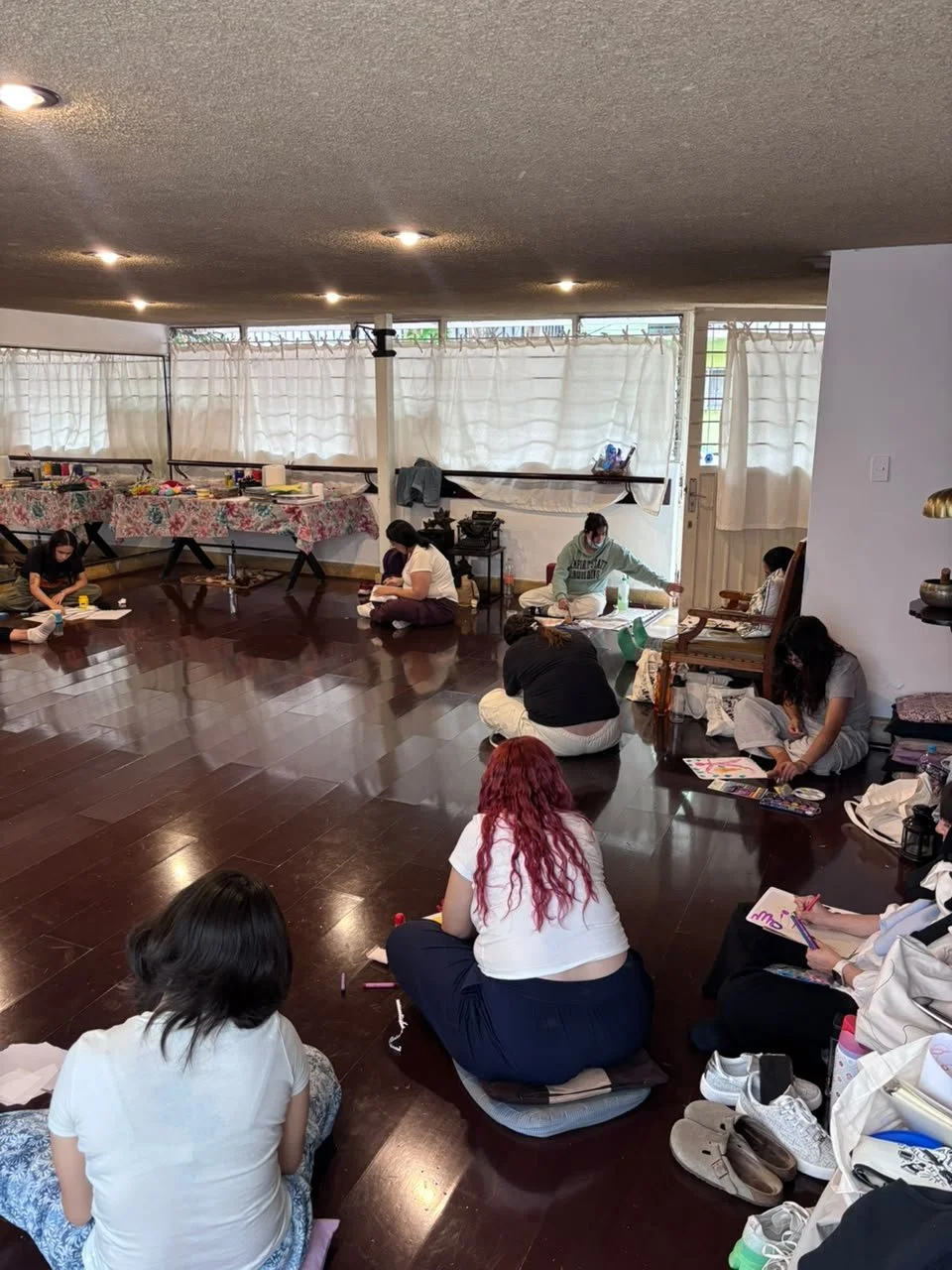 Grupo de personas sentadas en el suelo participando en una actividad creativa, en un espacio cerrado con cortinas blancas y piso de madera oscura.