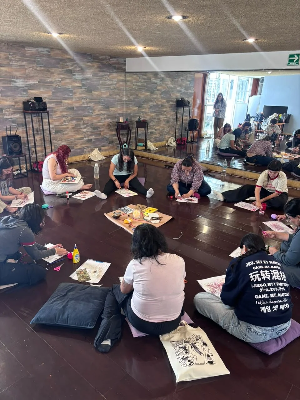 Grupo de personas sentadas en el suelo participando en una actividad de arte, con materiales y papeles frente a ellas en una habitación con pared de piedra y espejo grande.