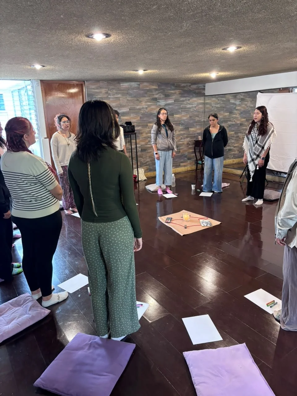Grupo de mujeres en una actividad interior, algunas de ellas de espaldas, participando en una reunión o taller con materiales y almohadones en el suelo.