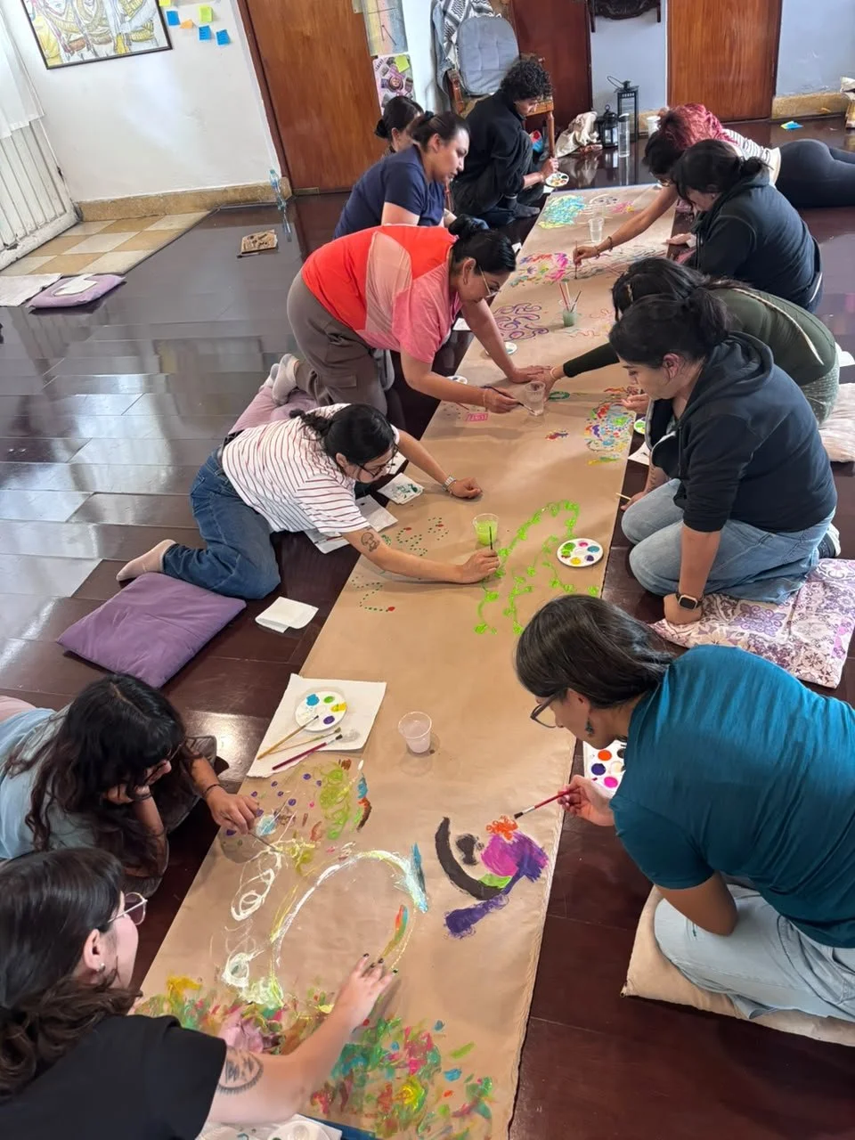 Grupo de personas pintando un amplio mural en el piso con acuarelas y pinturas acrílicas, usando pinceles y paletas de colores.