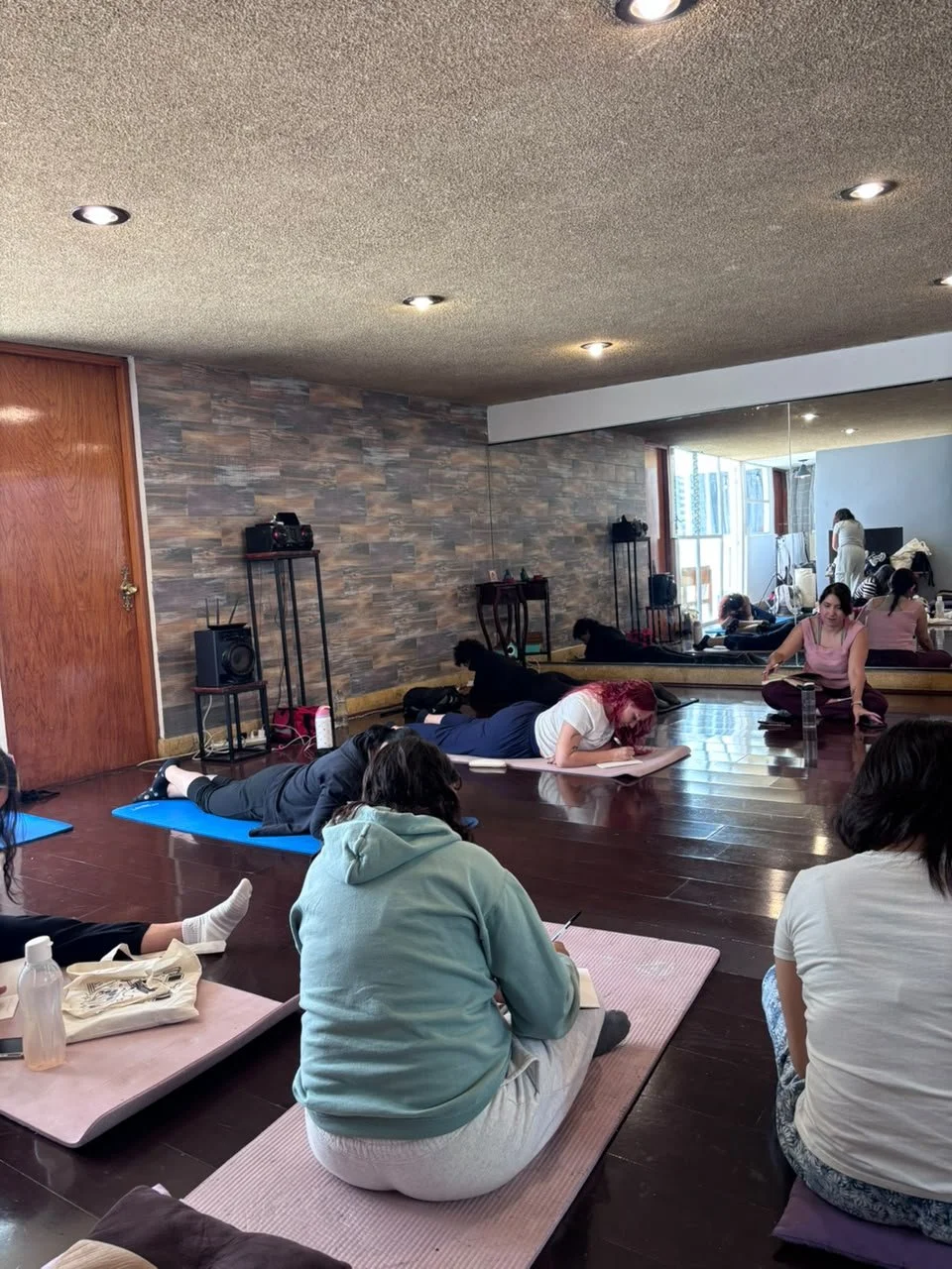 Varias personas en una clase de yoga o ejercicio en una sala con suelo de madera y paredes de piedra, algunas en posición de descanso o haciendo ejercicios