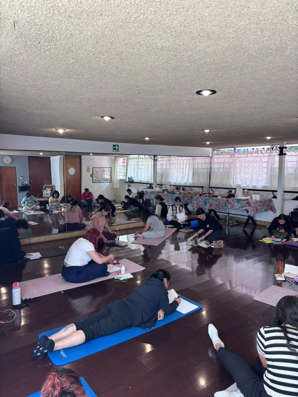 Personas en una clase de  arteterapia  o meditación en una sala con suelo de madera, algunas en colchonetas y otras en el piso, participando en una actividad grupal.