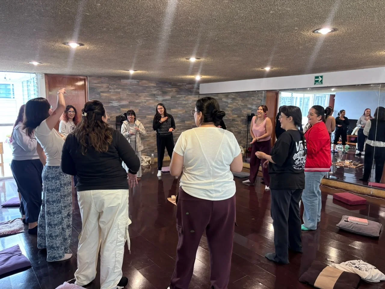 Grupo de mujeres participando en una actividad grupal en un espacio interior, algunos están de pie y otros sentados en colchonetas, en una actitud de interacción y conexión.