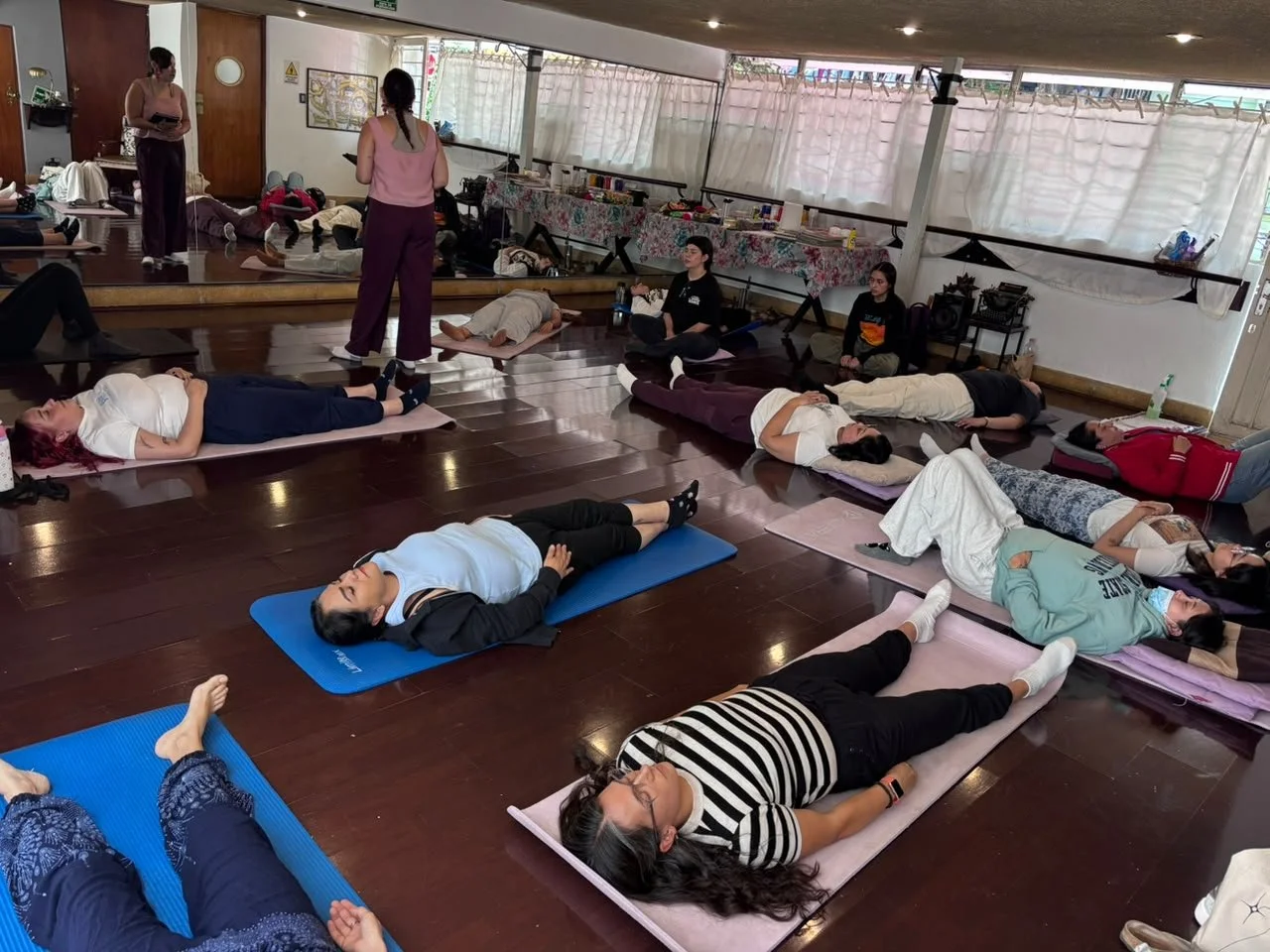 Personas participando en una clase de relajación o meditación en interiores, acostadas en colchonetas sobre un piso de madera, con algunas personas en posición de meditación y otras descansando.