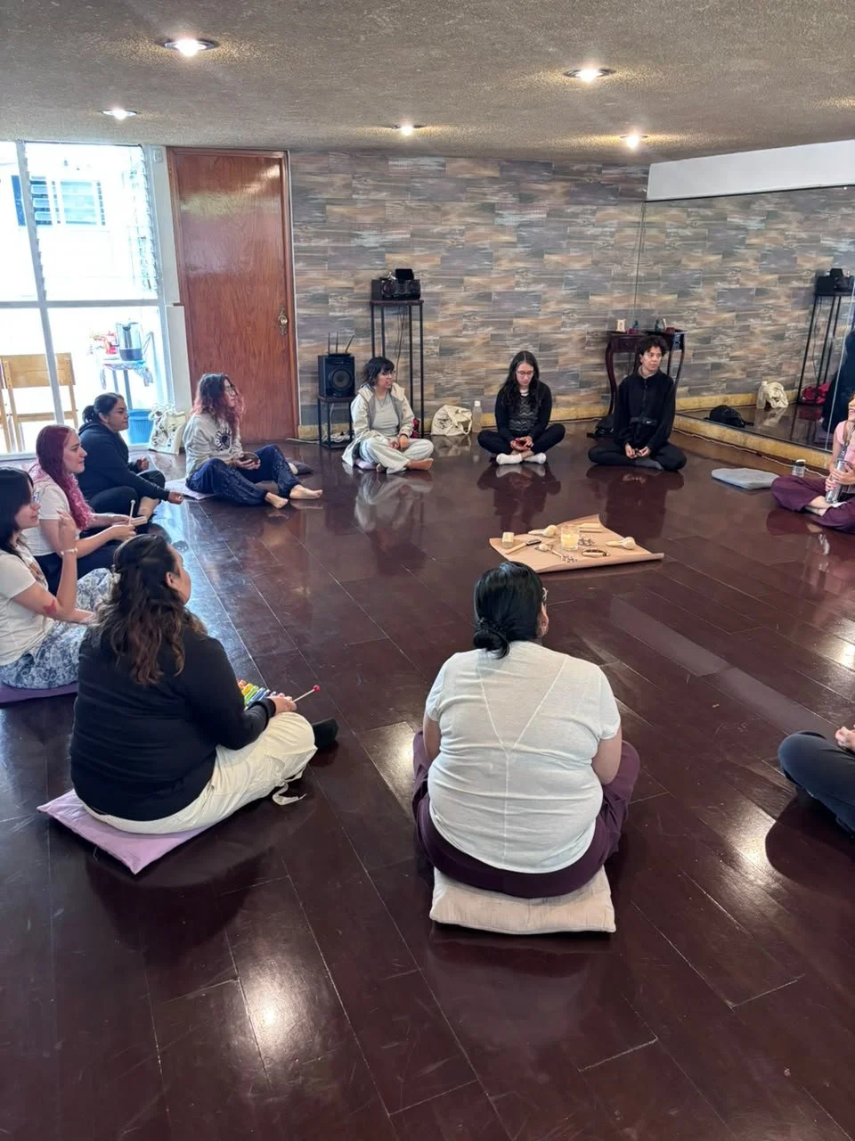 Personas sentadas en círculo en una sala, practicando meditación o yoga, con un altar en el centro con objetos decorativos y velas. Ambiente tranquilo con paredes de piedra y piso de madera.