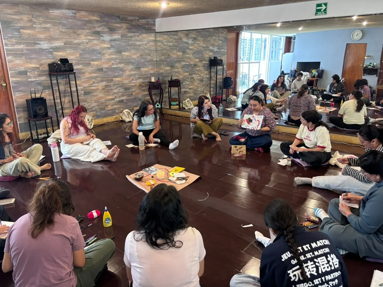 Grupo de personas sentadas en círculo en un taller o reunión en interiores con suelo de madera, algunas tomando notas y otras con materiales de arte o manualidades.