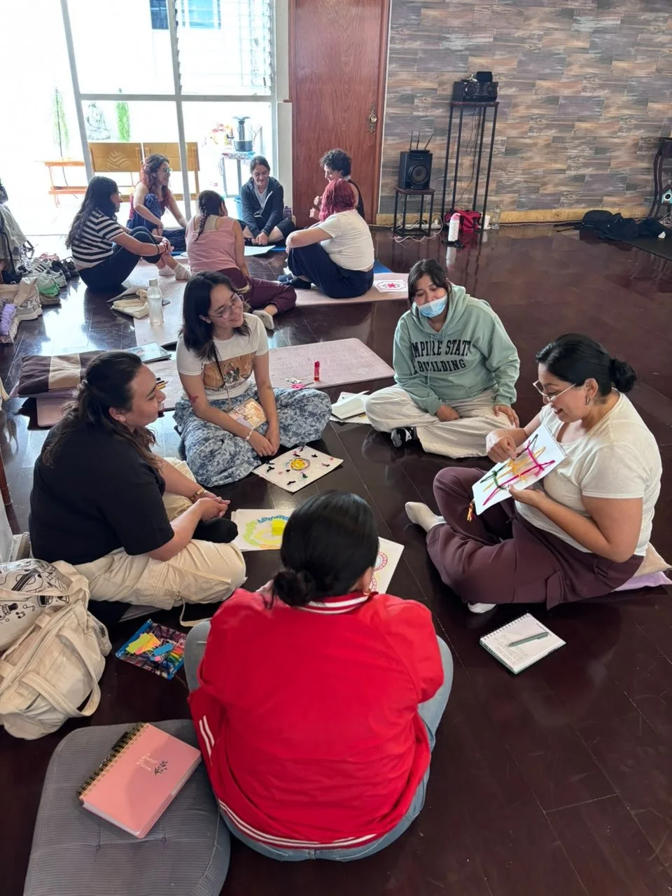 Grupo de personas sentadas en el suelo participando en una actividad creativa, algunas con papeles y colores, en un salón con madera en las paredes y ventanas grandes.