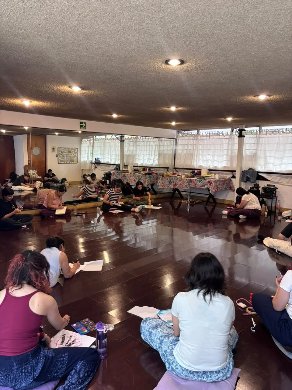 Grupo de personas sentadas en el suelo en un salón, participando en una clase o taller, con mesas y decoraciones en el fondo.