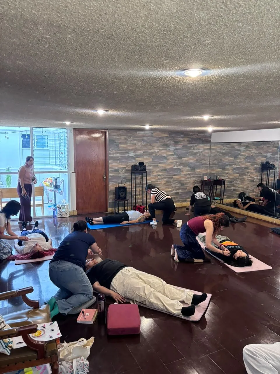 Grupo de personas en una clase de  arteterapia  o meditación en un salón con suelo de madera, algunos están acostados y otros en diferentes posiciones de relajación, con instructora guiándolos.