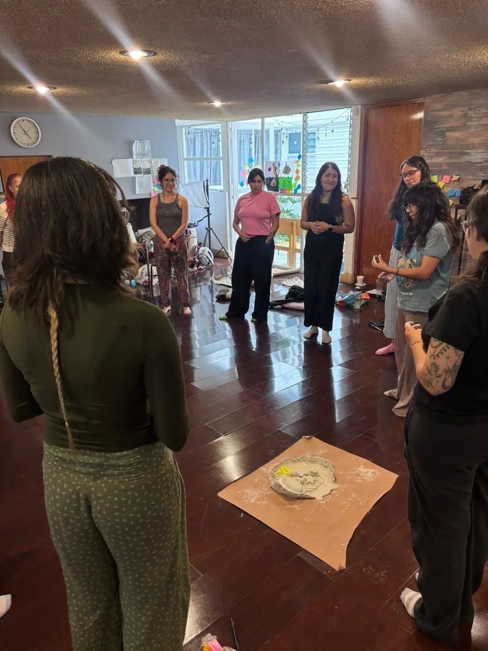 Grupo de mujeres en círculo en un espacio interior, con un horno de arcilla en el centro en una hoja de papel. 