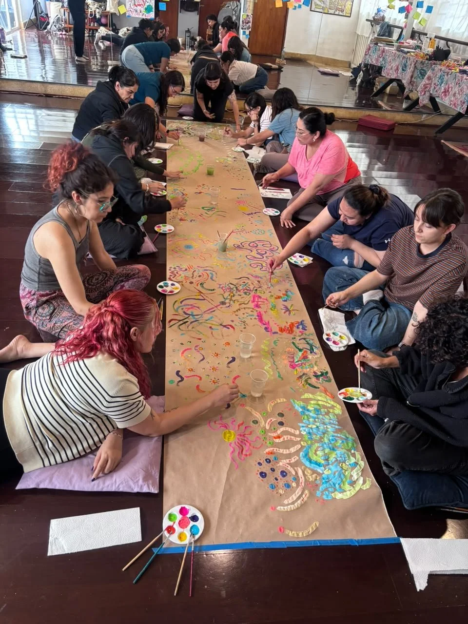 Varias personas pintando en una larga hoja de papel colocada en el suelo en un taller o clase. Están usando pinceles y pinturas de colores, sentadas en el suelo, en un ambiente decorado con dibujos y notas en las paredes.