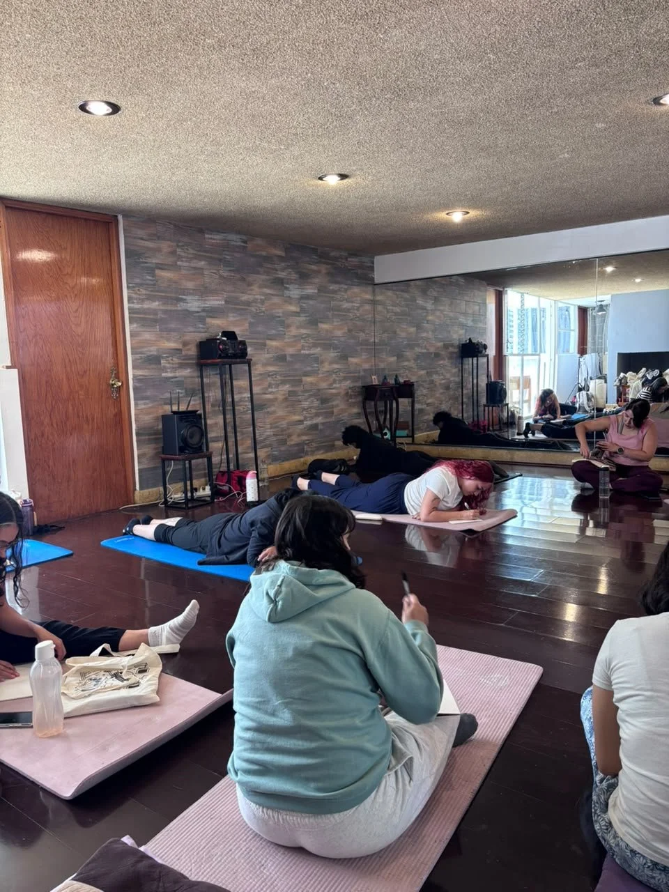 Varias personas en una clase de  arteterapia  o relajación en una sala con paredes de piedra y un espejo grande que refleja la escena.