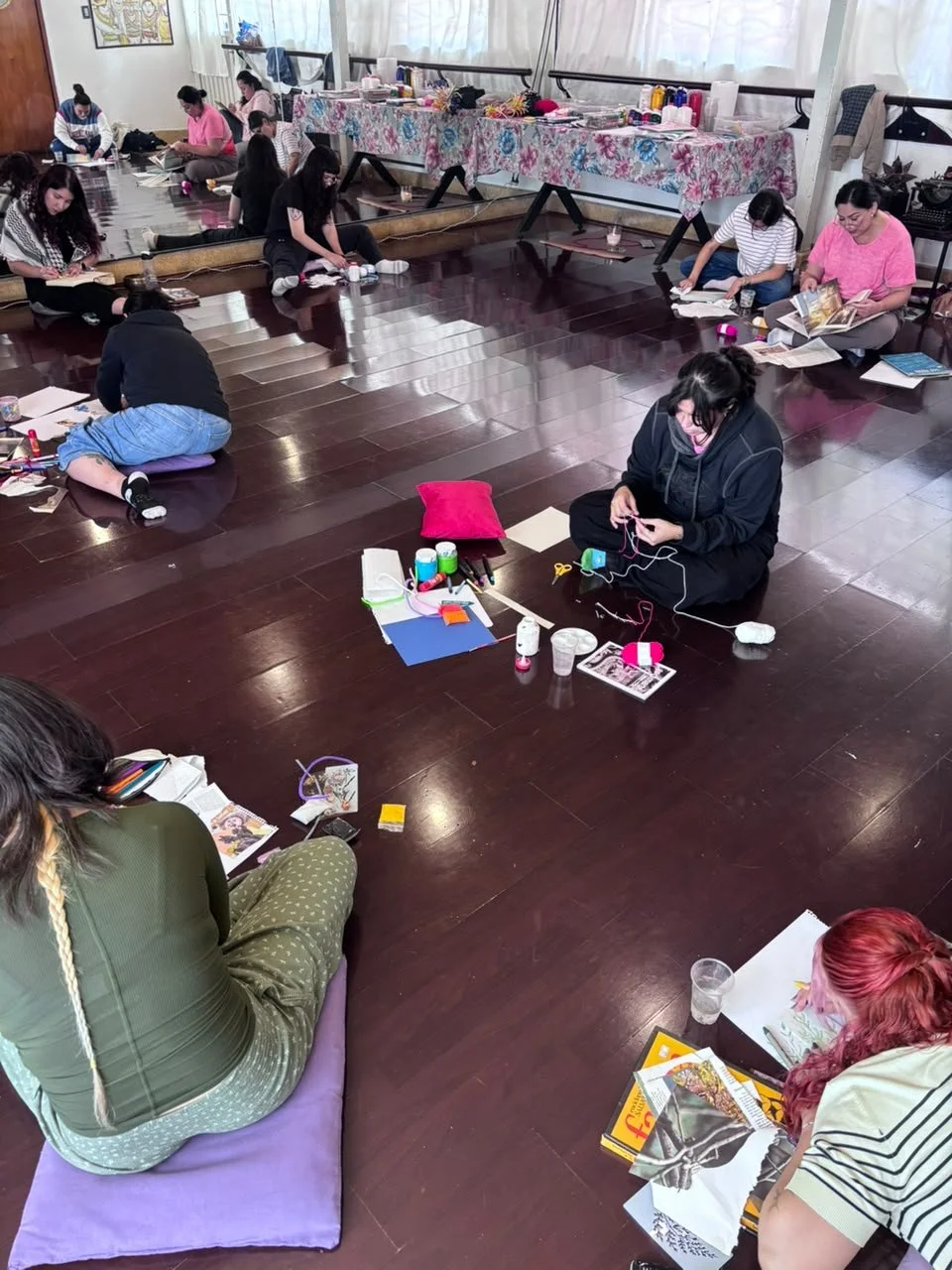 Grupo de personas sentadas en el suelo en una sala, participando en un taller o actividad artística, con libros, telas y materiales de manualidades.