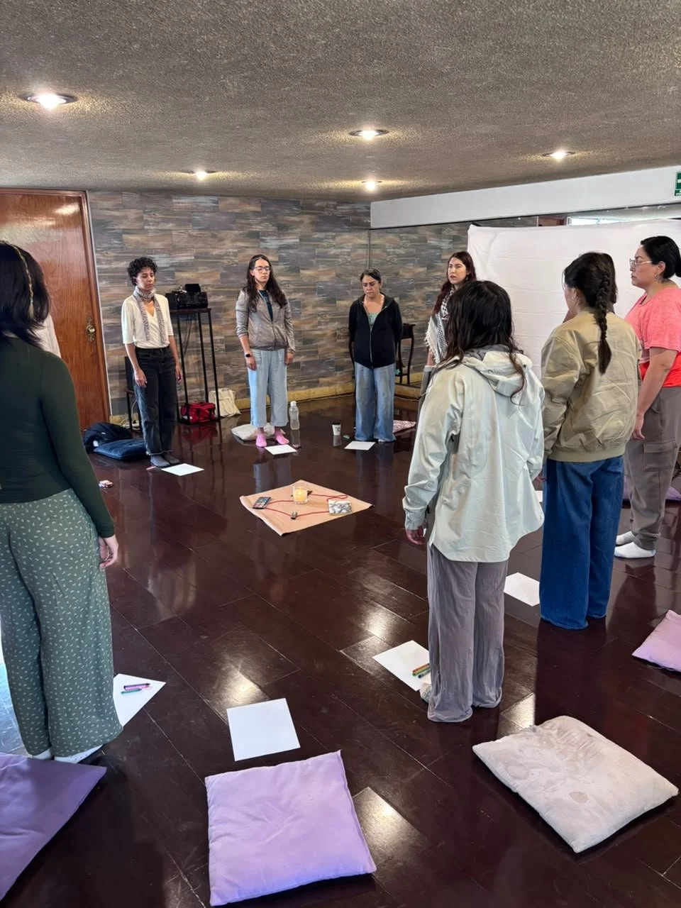 Grupo de mujeres participando en una actividad en interiores, con cojines en el piso y objetos en el centro, en un espacio con paredes de piedra y techo de acabado rincón.