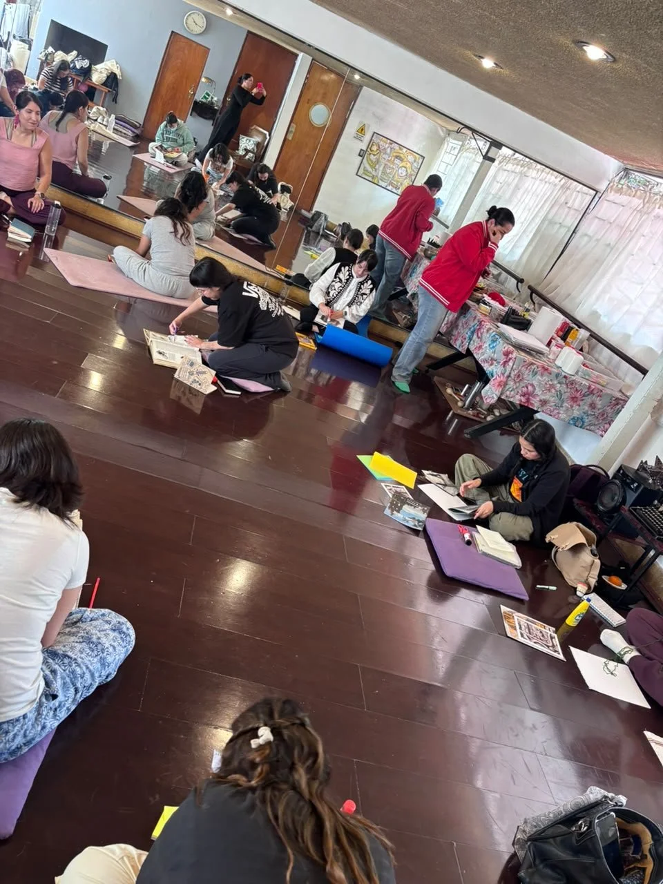 Grupo de personas participando en un taller de arte o artesanía en un salón con madera en el piso y paredes blancas, algunas con cortinas blancas y muebles con tejidos florales, varias están sentadas en el suelo y otras en una superficie elevada, alg