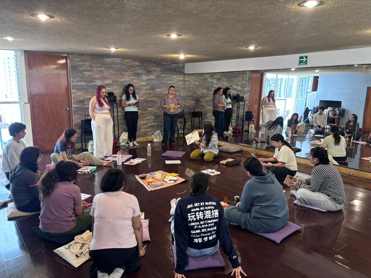 Grupo de personas participando en una actividad en una sala con piso de madera y paredes de piedra decorativa, sentadas en cojines en el suelo, con algunas frente a un espejo y otras de pie en una fila.