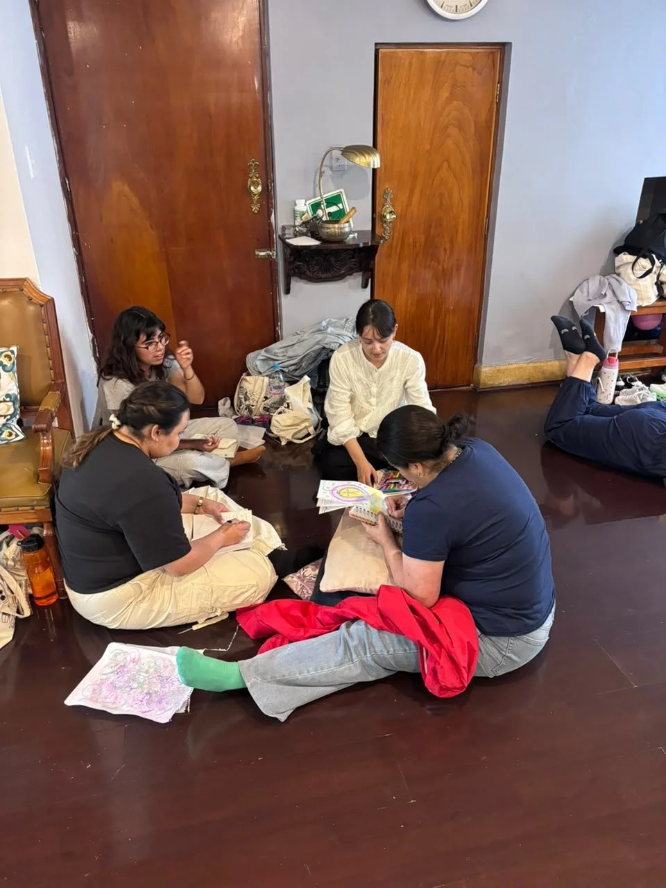 Cinco personas sentadas en el suelo en una habitación pequeña, concentradas en actividades creativas y de escritura, con objetos dispersos a su alrededor.