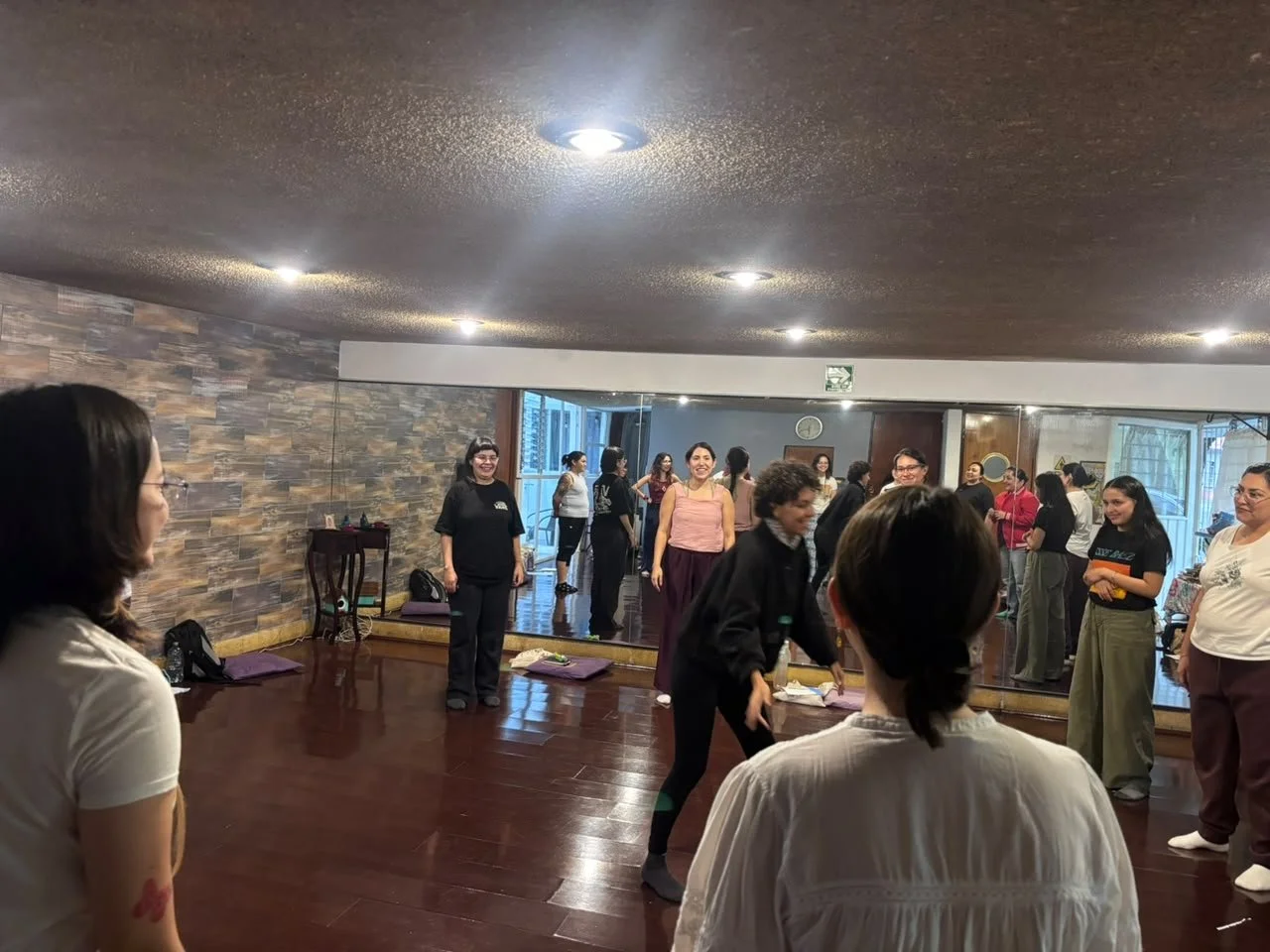 Grupo de personas en una clase de danza o ejercicio en un salón con pared de espejos y suelo de madera, participando y sonriendo.