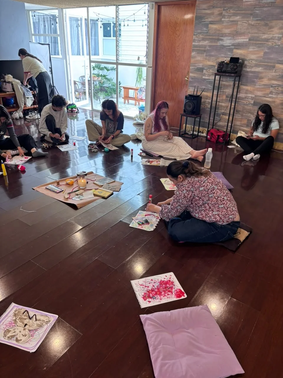 Personas en una habitación participando en una actividad artística, algunas están pintando en el suelo con pinturas y pinceles, en un ambiente que parece ser un taller o clase de arte, con una pared de madera y espacio abierto hacia una terraza con p