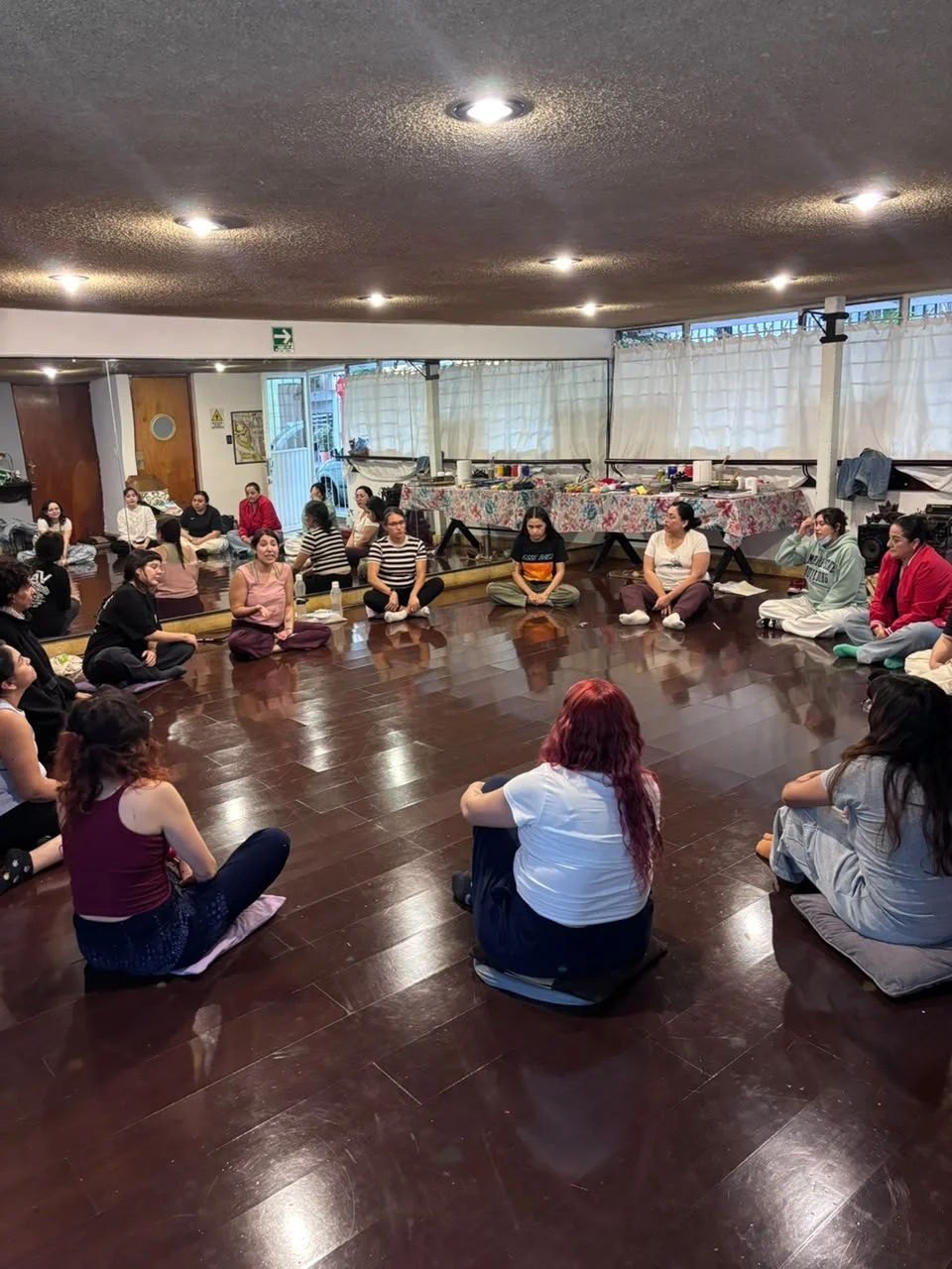 Un grupo de personas sentadas en círculo en el suelo de una sala, participando en una actividad grupal, con luz amarilla desde el techo y cortinas blancas en las ventanas.