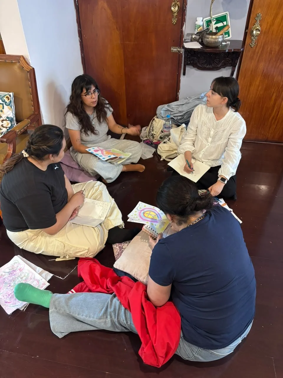 Cuatro mujeres sentadas en un piso de madera, conversando y compartiendo dibujos y notas. Una de ellas tiene una libreta y otra una pluma. Hay materiales artesanales y bolsas alrededor, en un espacio interior con puertas de madera y decoración vintag