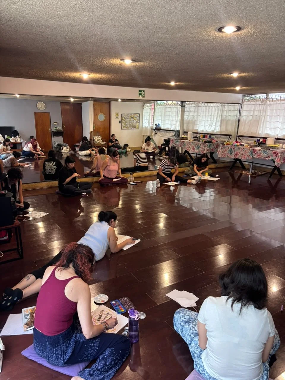 Grupo de personas participando en una clase de arte o manualidades, sentadas en el suelo con materiales de pintura y dibujo en un espacio interior