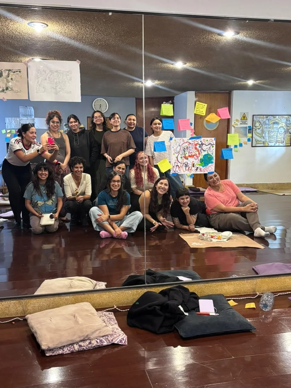 Grupo de personas posando para una selfie en un estudio con paredes azules y suelo de madera, reflexionando sobre una actividad artística, con materiales de arte como papel, dibujos y notas adhesivas en la pared.