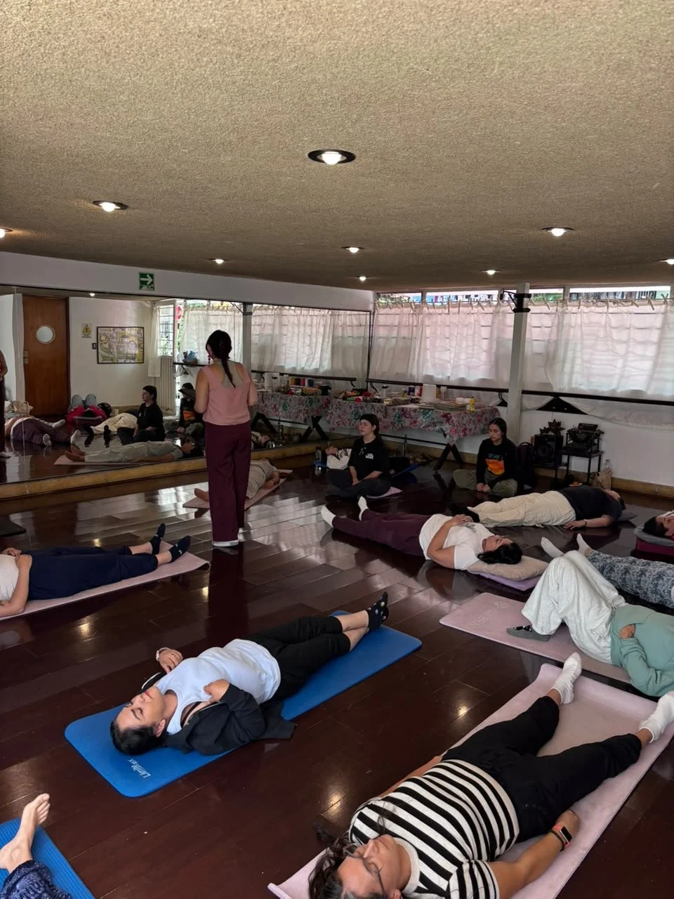 Grupo de personas practicando yoga en un estudio con suelo de madera y cortinas blancas, una instructor dando instrucciones y algunos participantes con colchonetas de yoga.