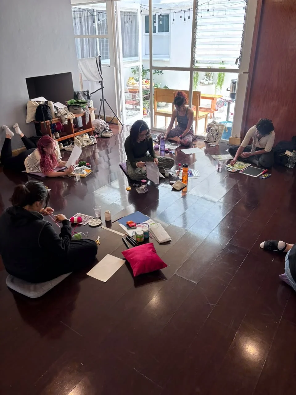 Grupo de cinco personas sentadas y acostadas en el piso de una habitación, trabajando en proyectos con materiales artísticos. Una ventana grande da al exterior con muebles y plantas