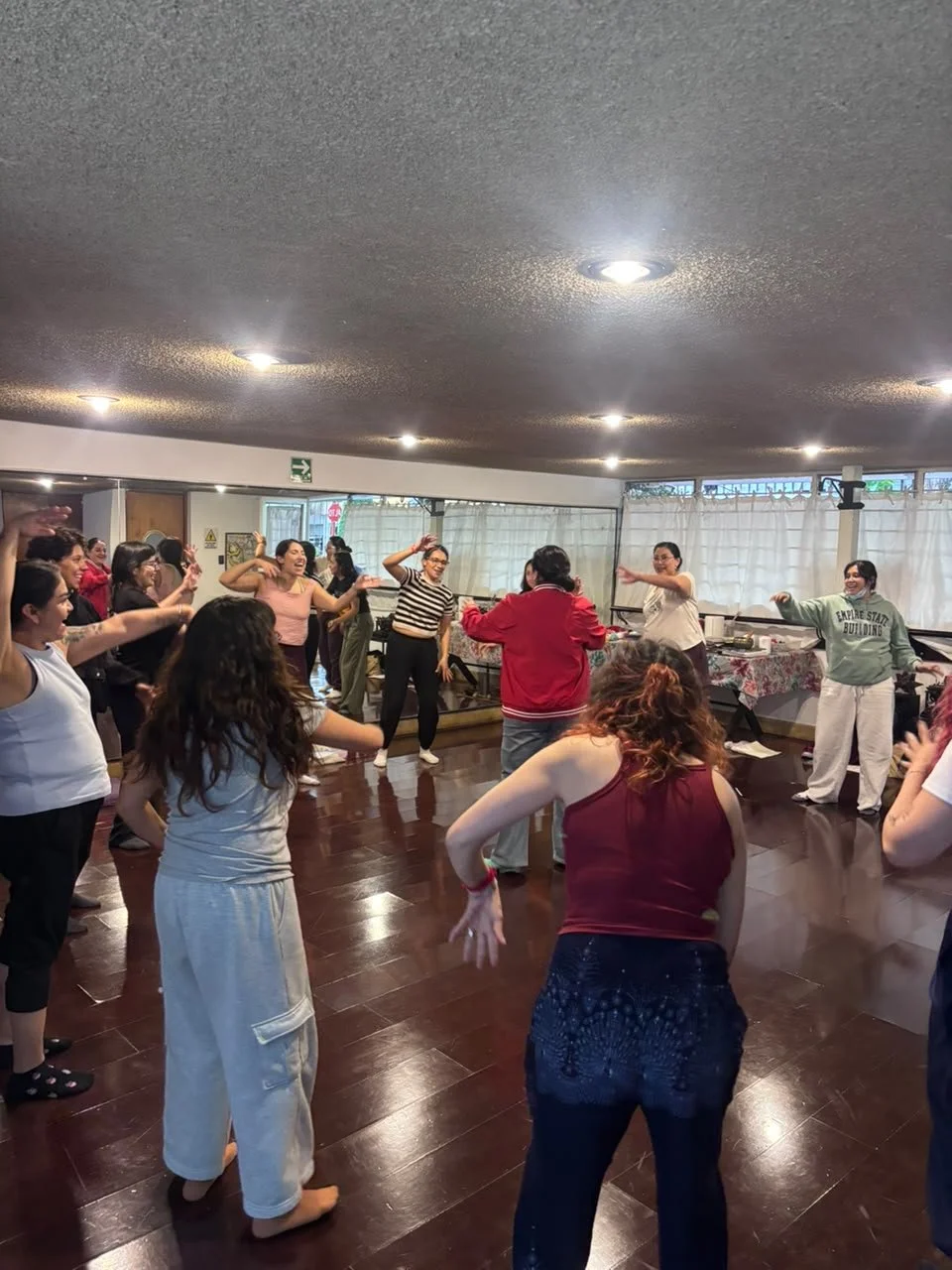 Grupo de mujeres bailando y disfrutando en una sala con suelo de madera y cortinas blancas.