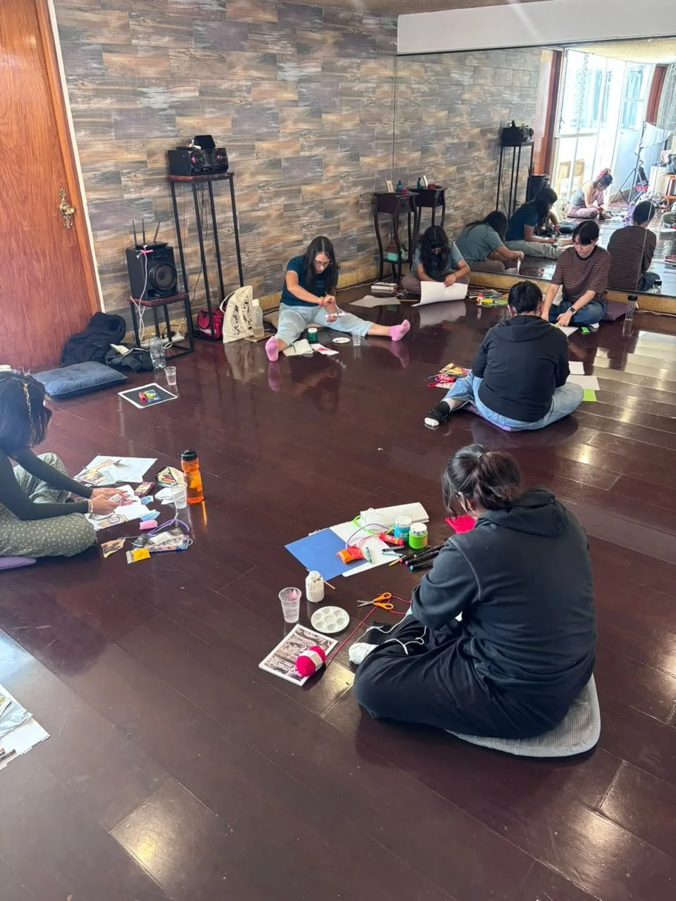 Grupo de personas sentadas en el suelo en una reunión o taller, con materiales de manualidades y notebooks, en una habitación con pared de piedra y pared de espejo.