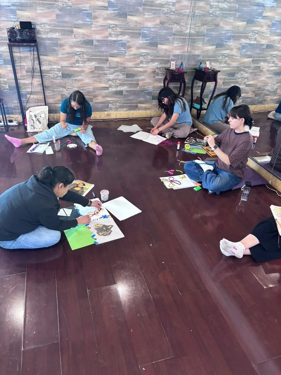 Varias personas sentadas en el suelo haciendo manualidades, con materiales y papeles, en una habitación con paredes de piedra y piso de madera.