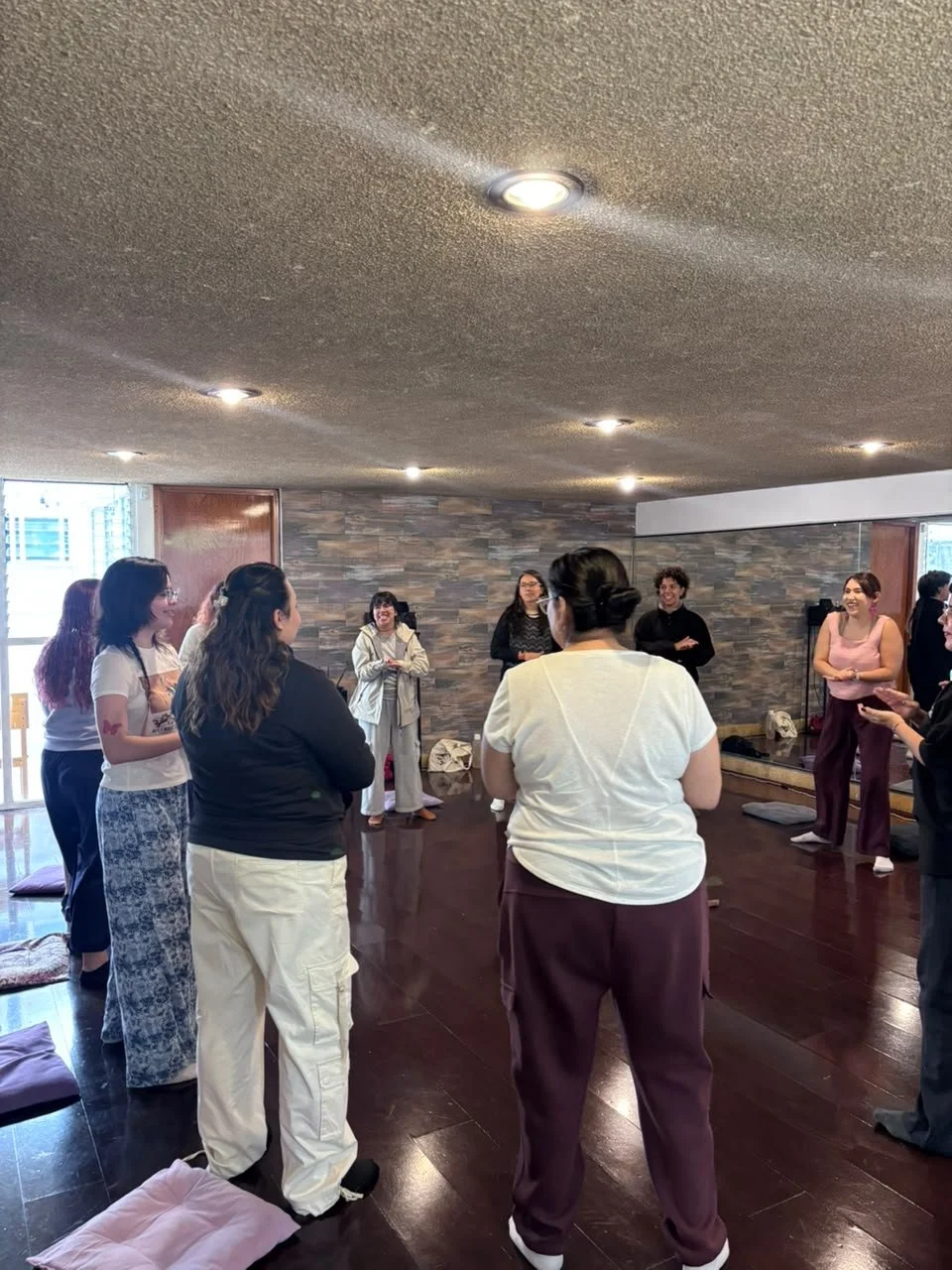 Varios adultos en círculo en una sala de yoga o meditación, algunos con ropa cómoda, en un ambiente con pared de piedra y piso de madera, practicando una actividad grupal.