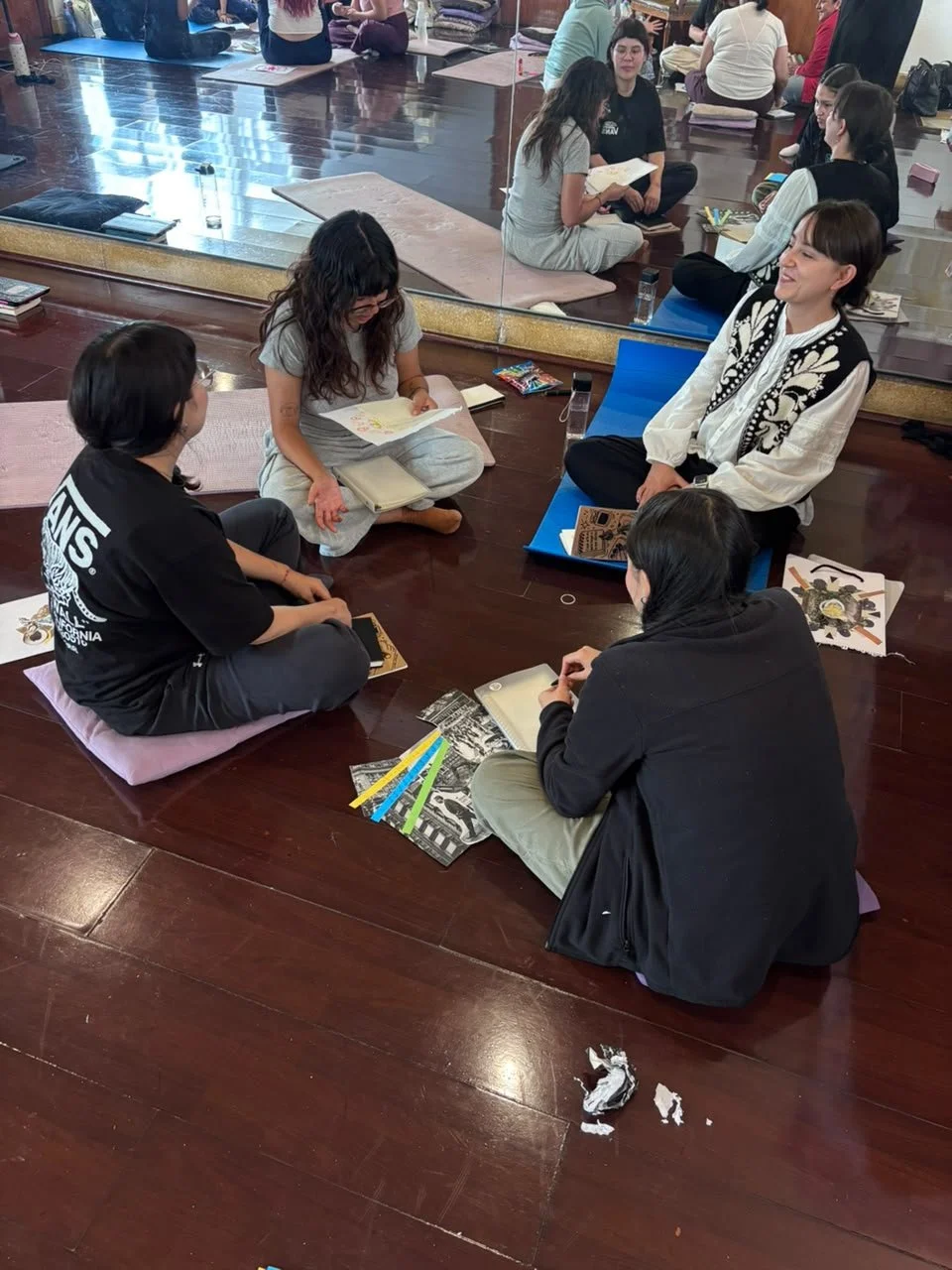 Grupo de personas sentadas en el suelo, participando en una actividad de lectura o discusión en un espacio interior, con libros y papeles esparcidos alrededor.