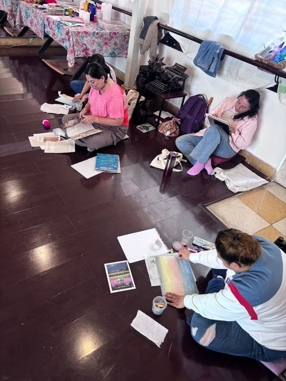Cuatro personas sentadas en el suelo de una habitación, con materiales de arte y libros, participando en una actividad creativa.