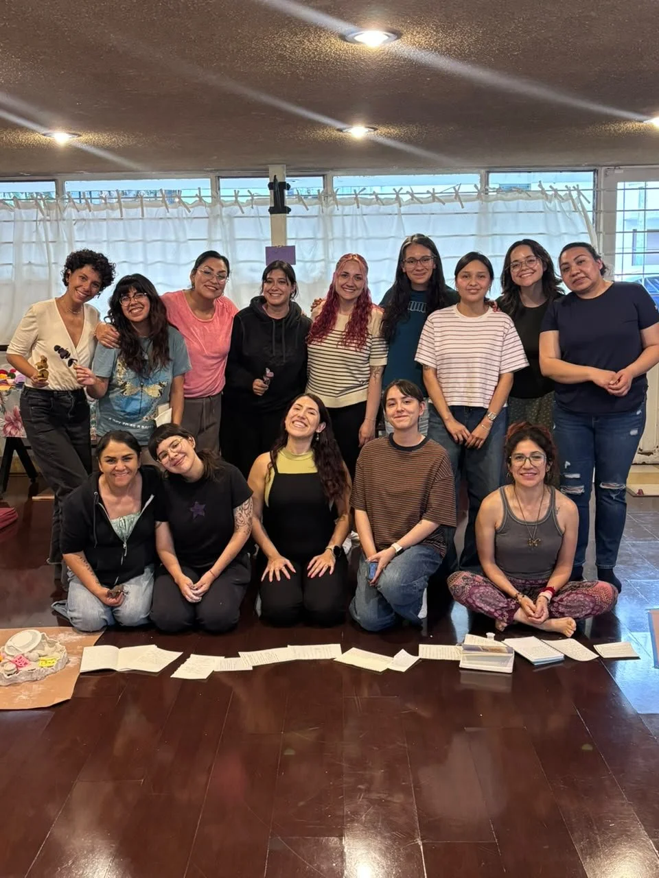 Un grupo de quince personas, en su mayoría mujeres, posando juntas en una habitación con cortinas blancas y ventanas grandes, todas sonriendo y mostrando un ambiente de celebración o reunión social.