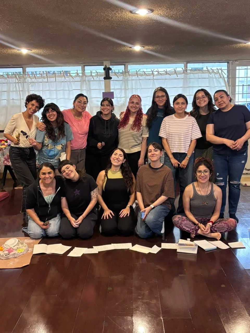 Grupo de veinte personas, en su mayoría mujeres jóvenes, posando juntas en un ambiente interior, algunos están de pie y otros sentados en el suelo, con risas y expresiones alegres.