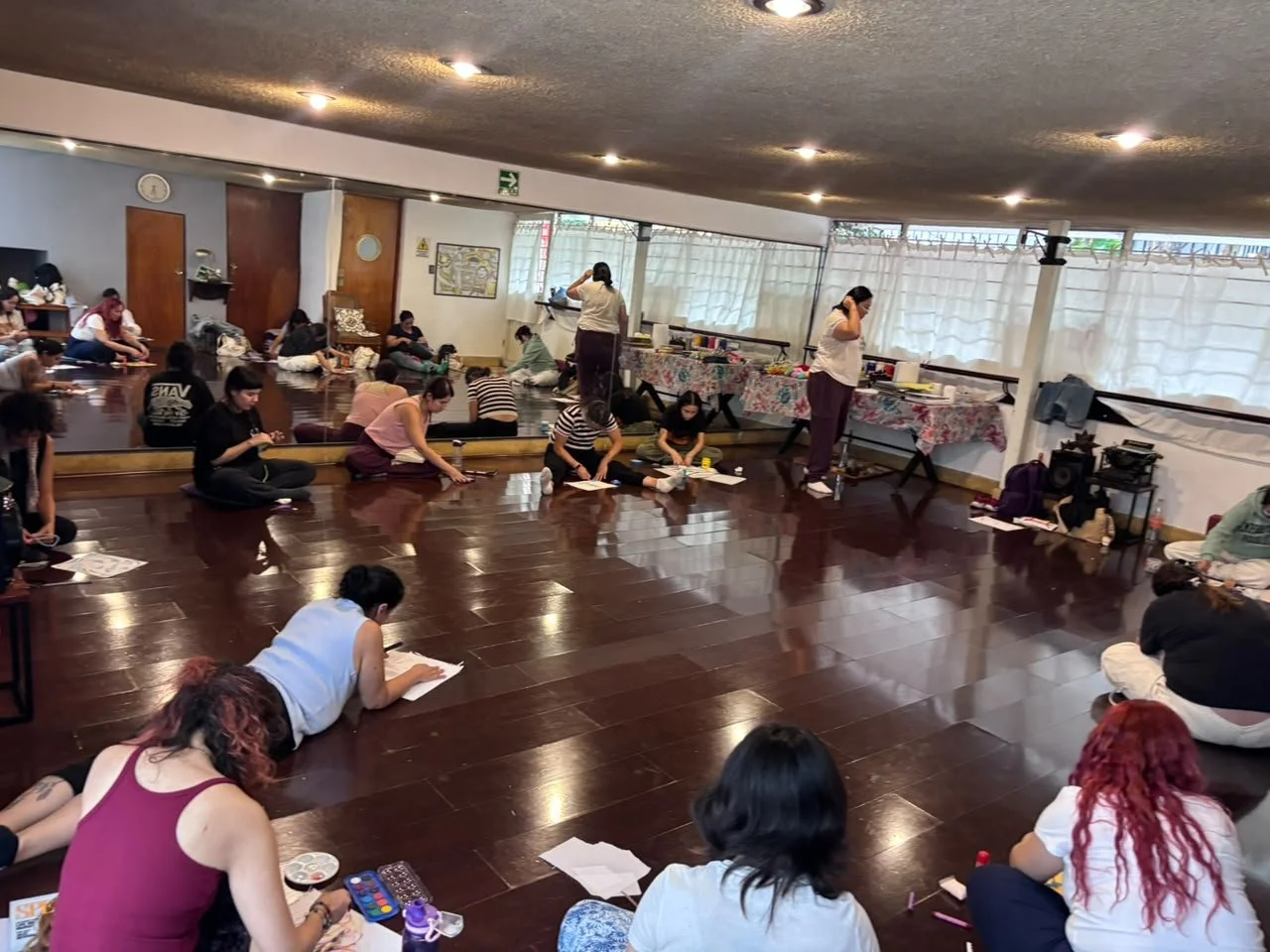 Grupo de personas sentadas en un piso de madera, participando en una clase o taller en un salón con paredes blancas y cortinas blancas. Algunas personas están tomando notas y otras observando.