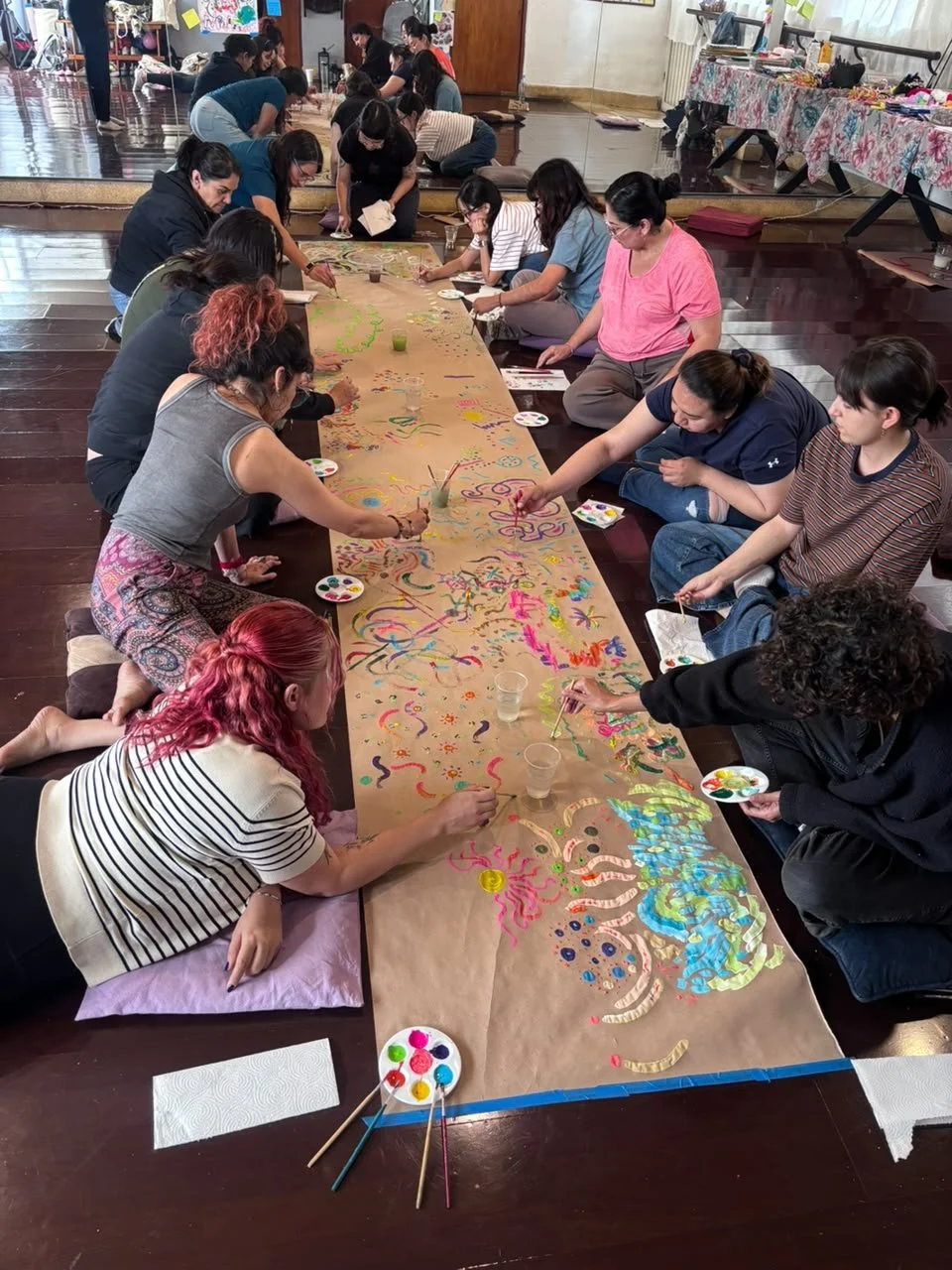 Grupo de personas pintando un mural largo en papel kraft en el suelo, en un taller o clase. Varias paletas de pintura y pinceles en la mesa y en las manos de los participantes.