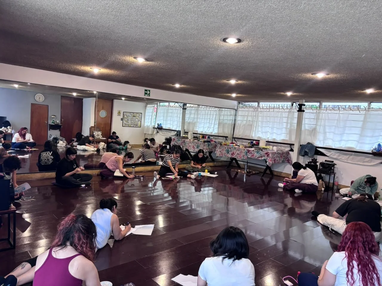 Grupo de personas en un salón de clases de yoga, muchas en posición de meditación, algunas en el suelo con notas o libros.