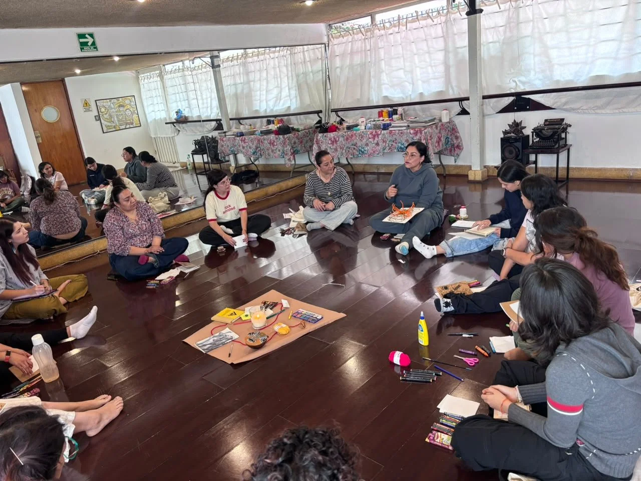 Grupo de personas sentadas en círculo en una sala, participando en una reunión o taller, con materiales y objetos en el centro.