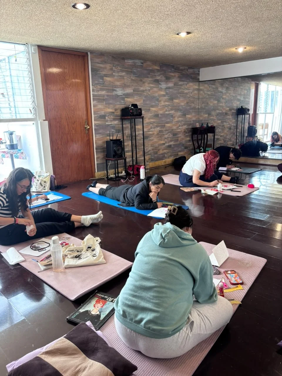 Varias personas en una clase de yoga o meditación en interior, en posición de loto, en un espacio con piso de madera y paredes con acabados de piedra, usando ropa cómoda y estirándose, con accesorios y materiales de estudio o ejercicio alrededor.