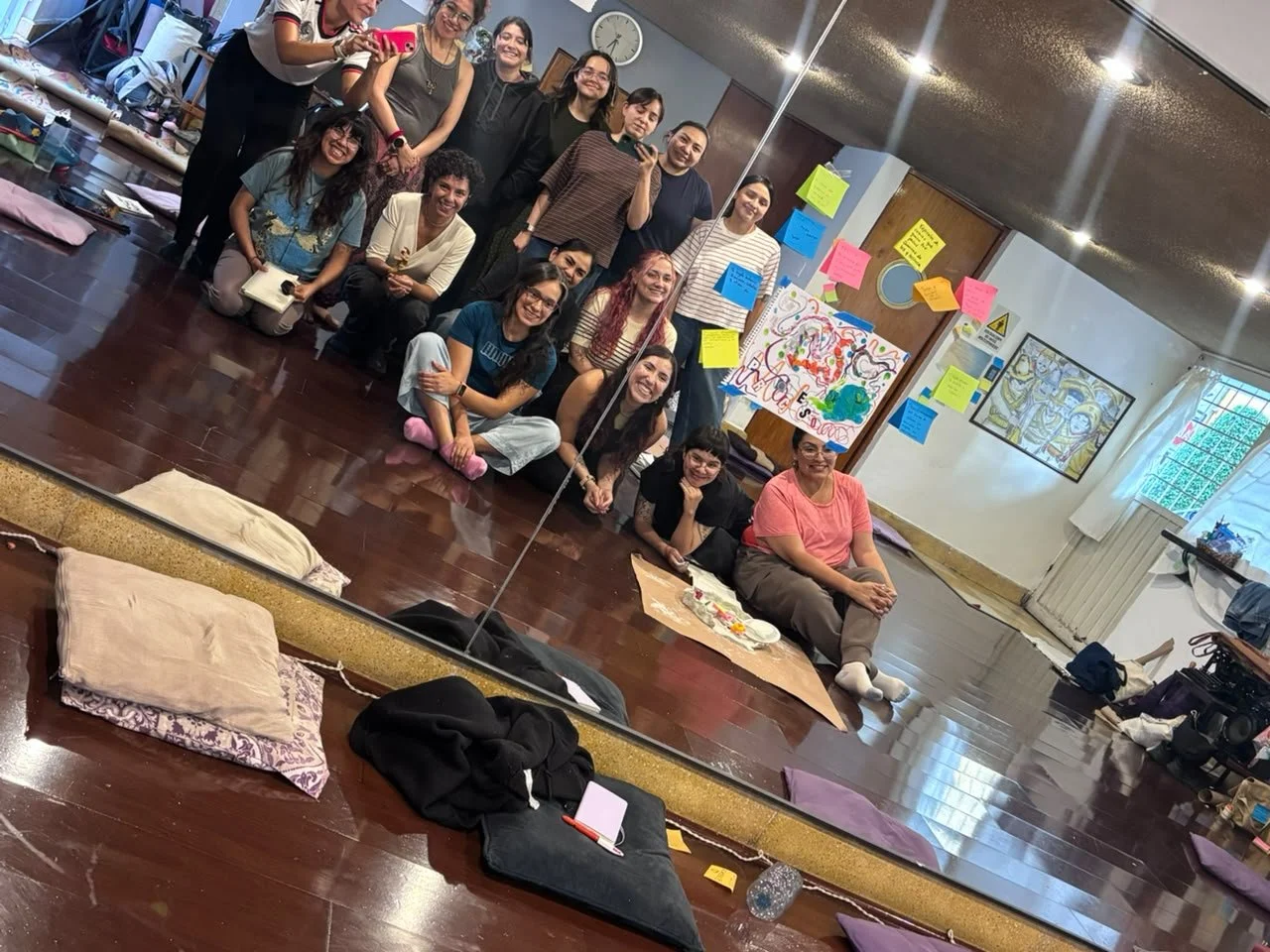Grupo de personas en una clase o taller creativo, posando frente a un espejo, con materiales y arte en el fondo.