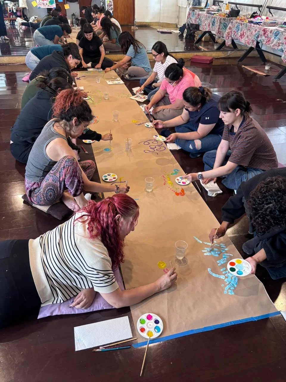Grupo de personas sentadas en el suelo, pintando en una larga hoja de papel, en un ambiente interior.