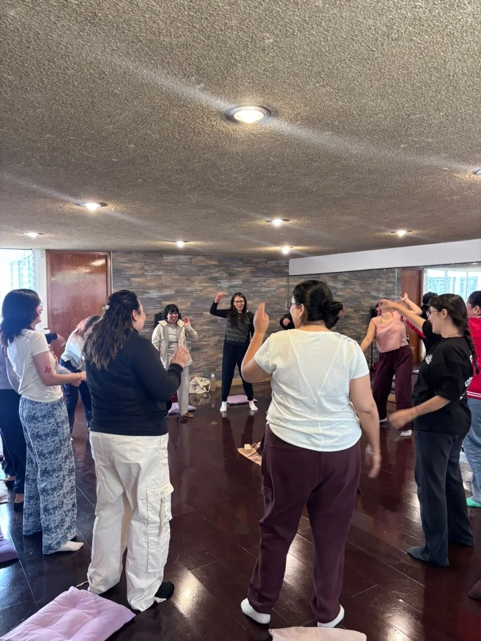 Grupo de personas participando en una actividad grupal en interiores, con una mujer en el centro levantando el brazo como si dirigiera o motivara, en un espacio con paredes de piedra y piso de madera.